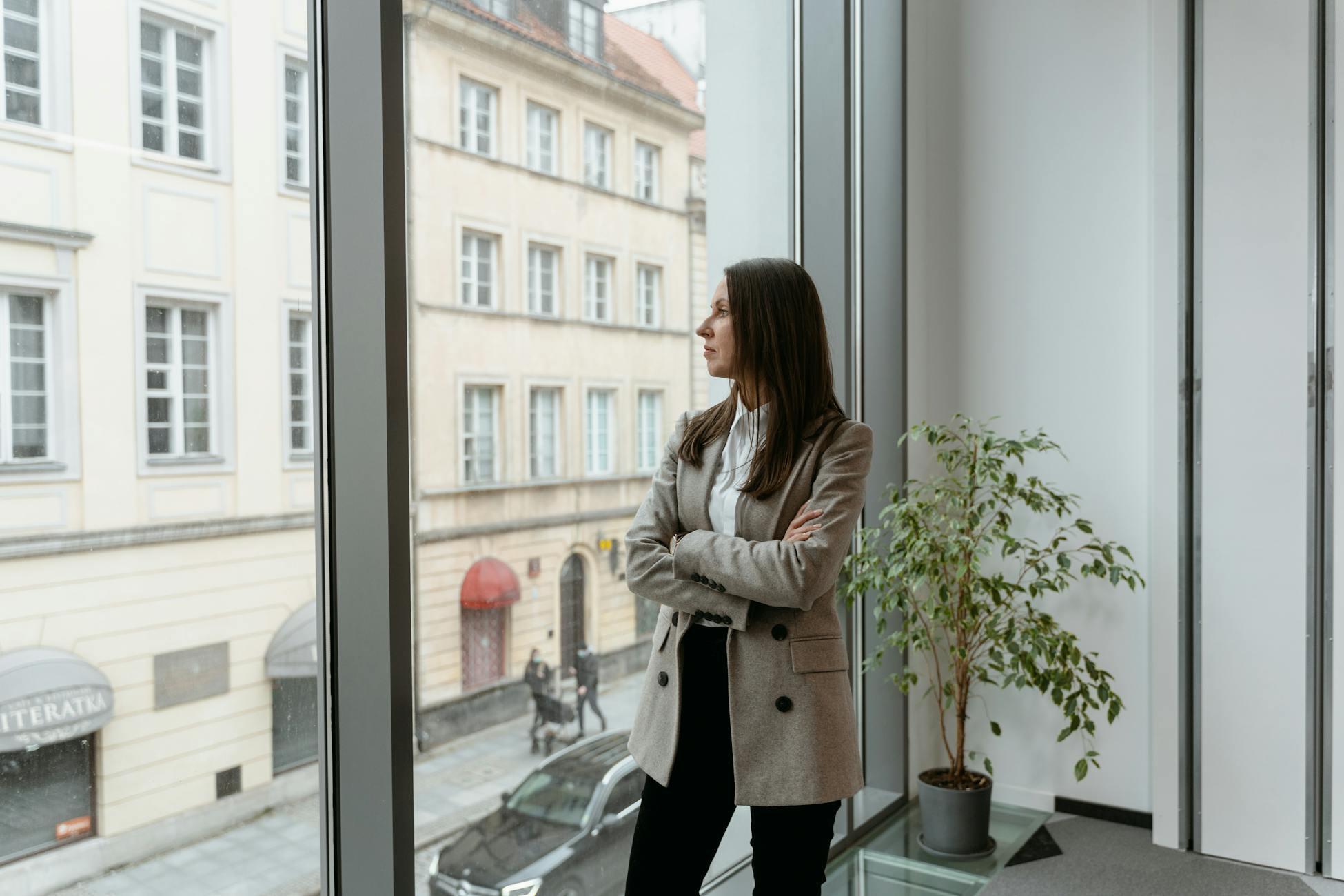 A woman in a blazer stands by a large window, gazing at the cityscape from an office.