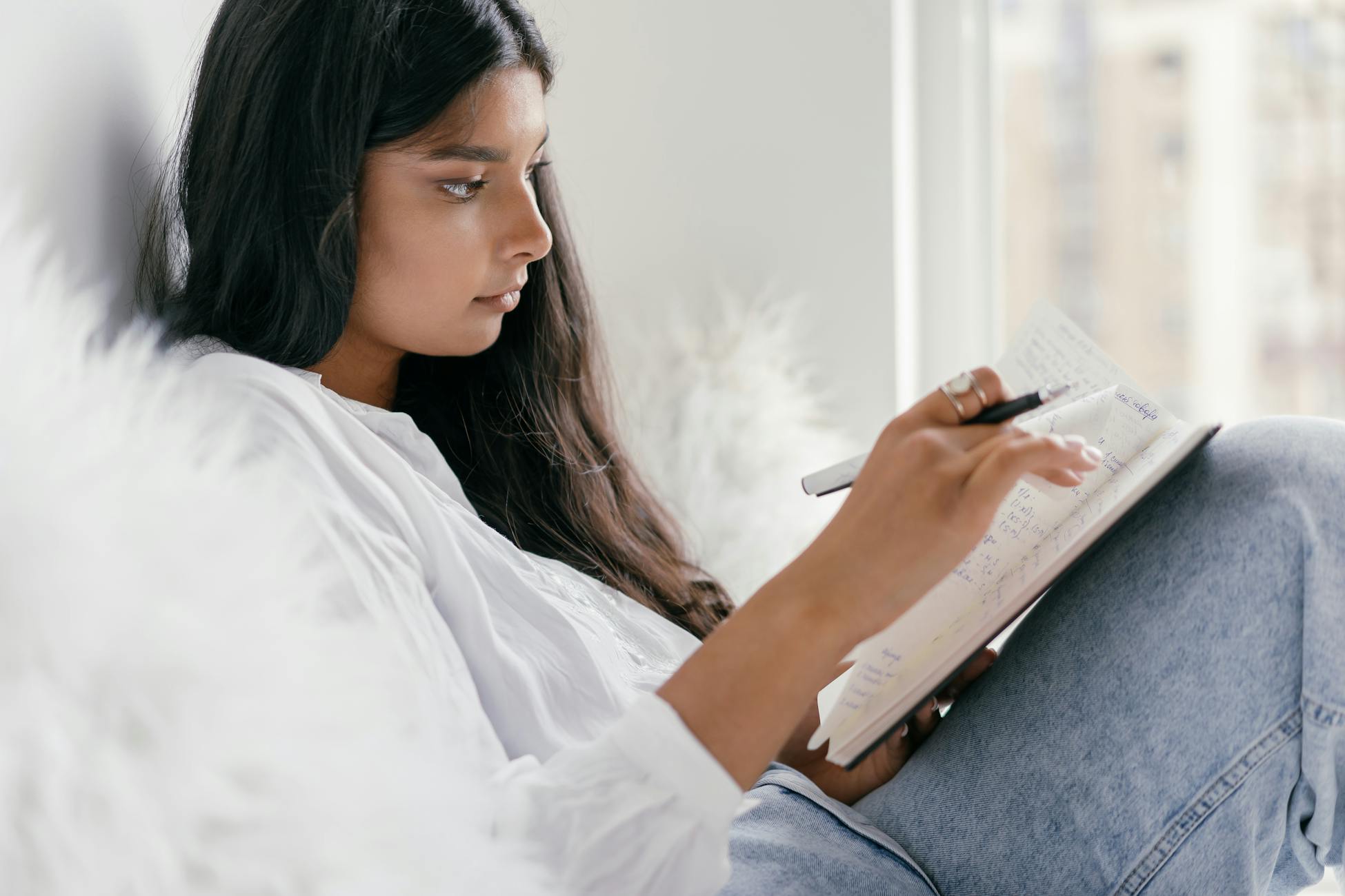 Young woman journaling indoors with a city view, surrounded by cozy pillows.
