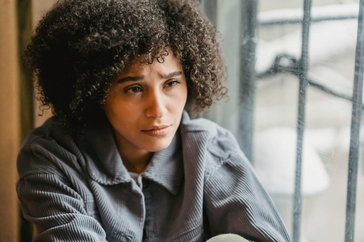 A young woman with curly hair sits pensively by a window, reflecting emotions indoors.
