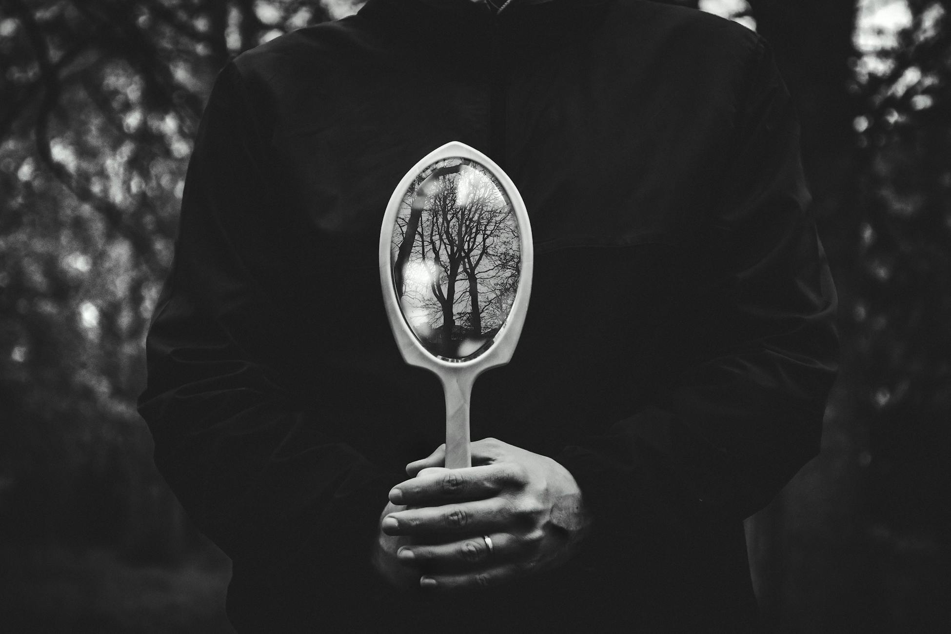 A person holding a mirror reflecting bare trees in a black and white setting.