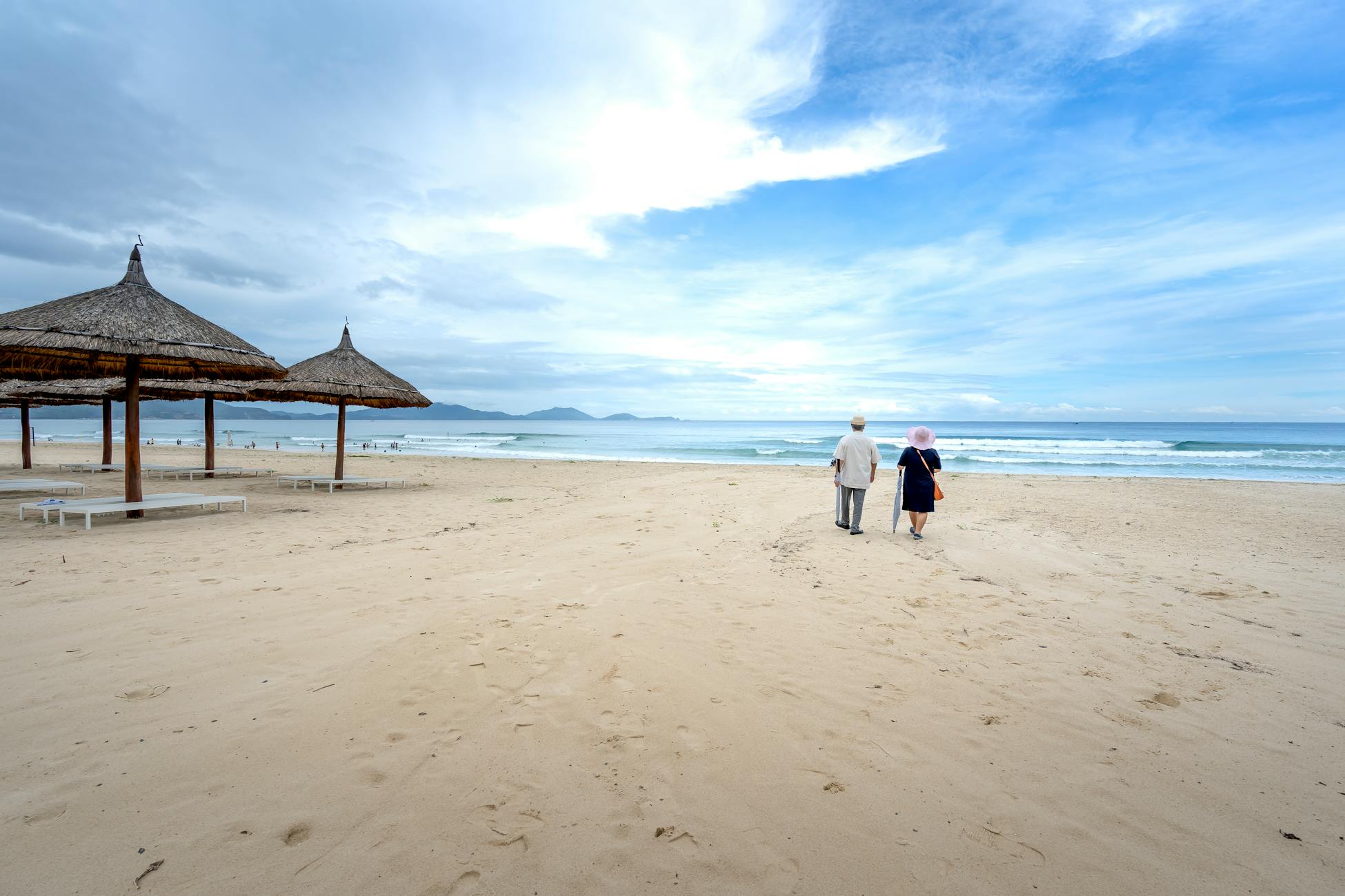 A peaceful scene of an elderly couple walking on a sandy beach under a partly cloudy sky.