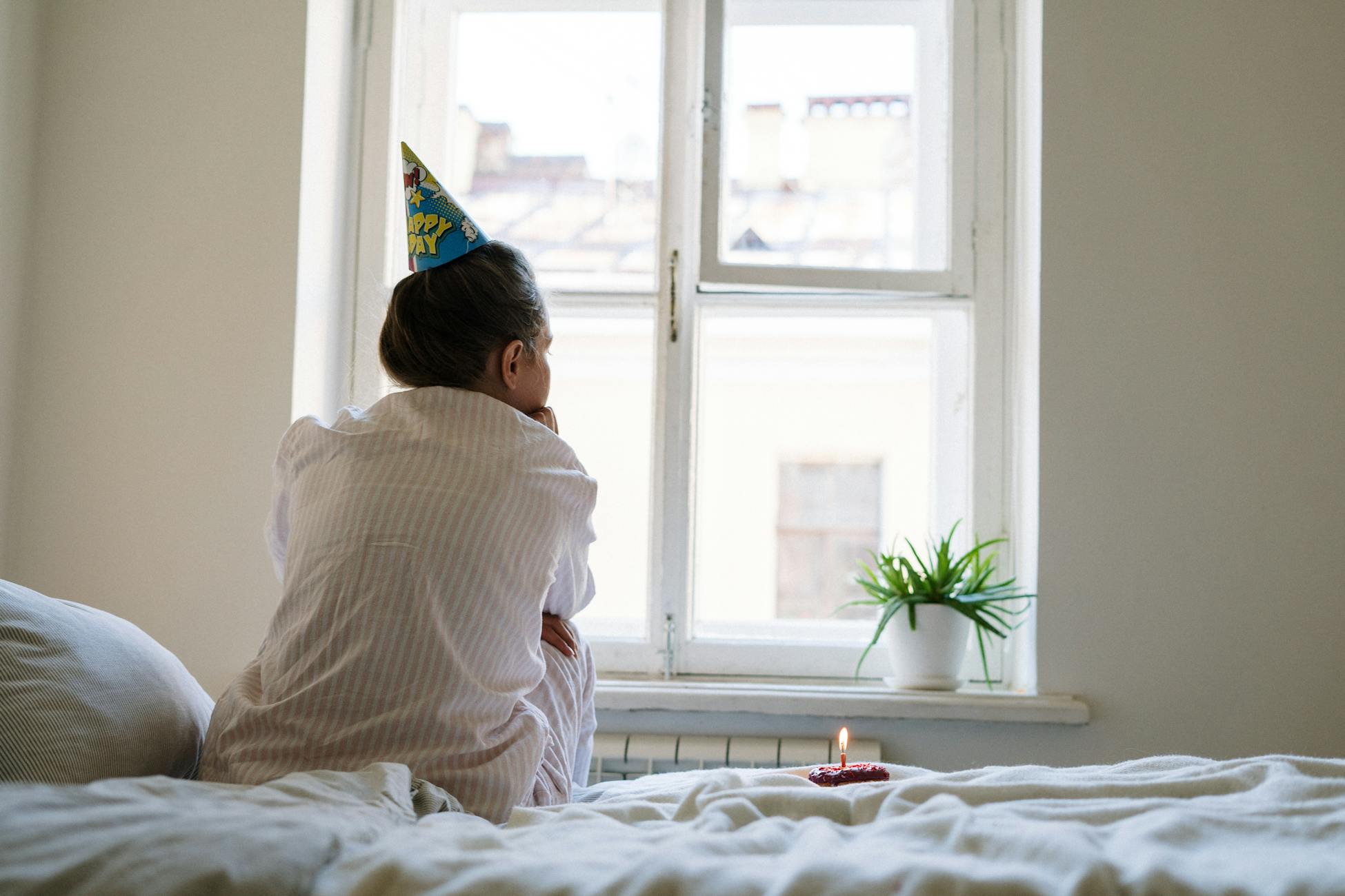 A person sits alone with a party hat and candle in a cozy bedroom, reflecting a quiet birthday moment.