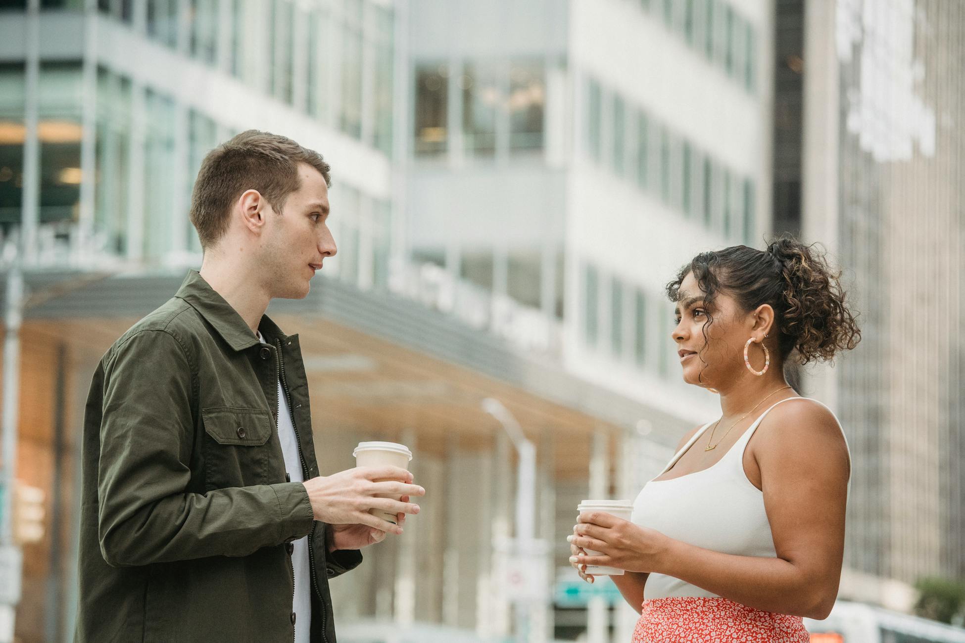 Side view of concentrated young multiracial male and female colleagues standing on street near modern skyscrapers and chatting with cups of takeaway coffee in hands