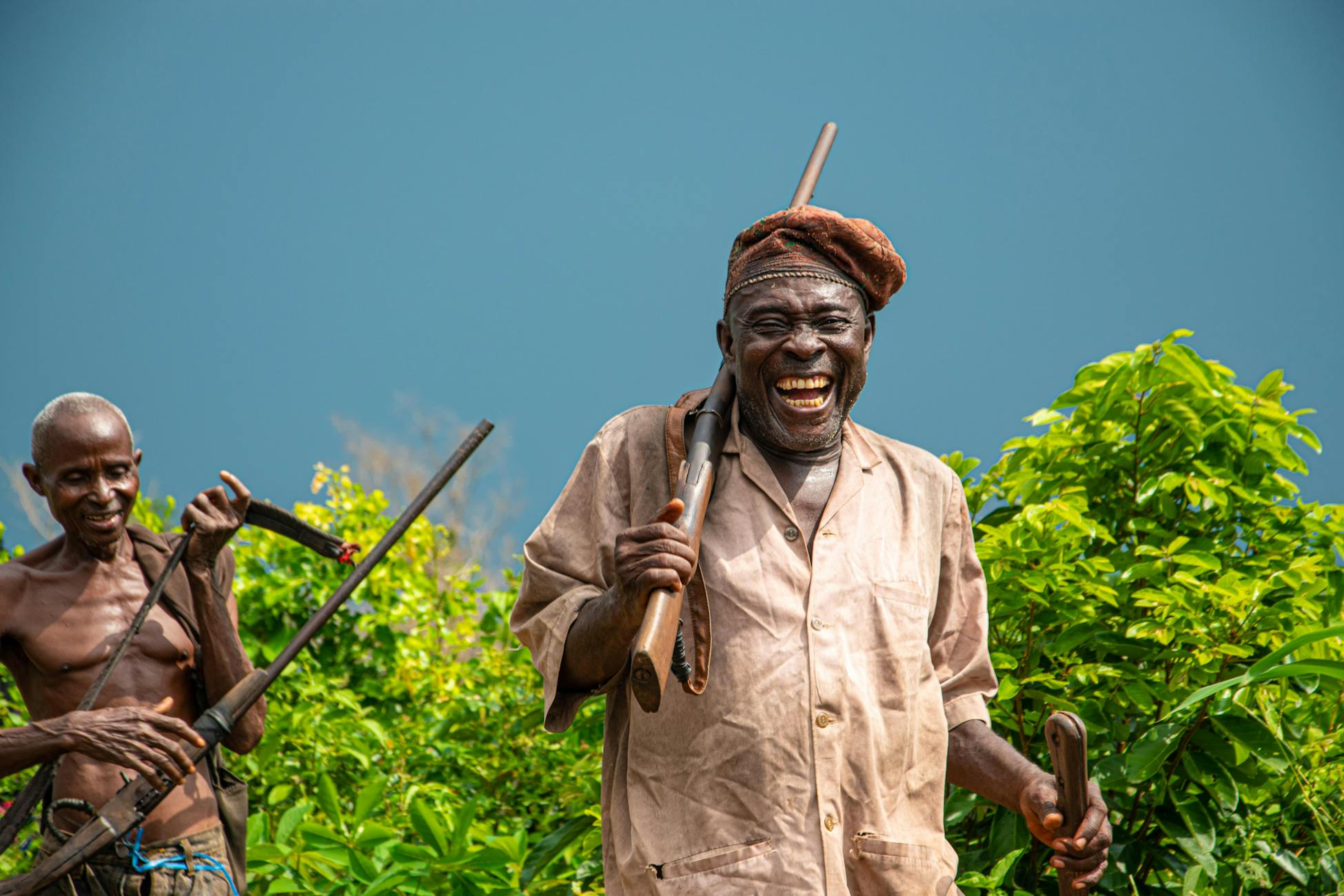 Two African men laughing and carrying rifles outdoors, exuding happiness and camaraderie.