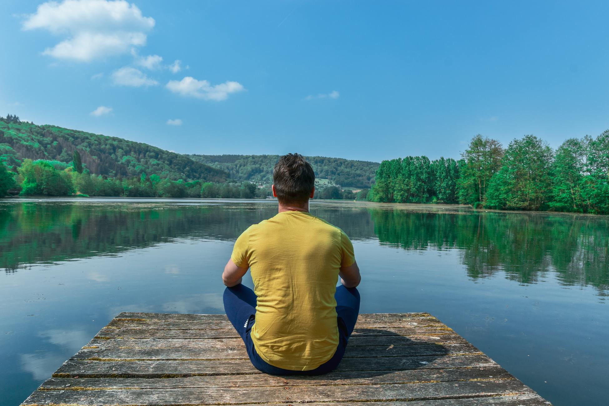 Man meditating on a peaceful dock by a serene lake in a lush landscape under a clear sky.