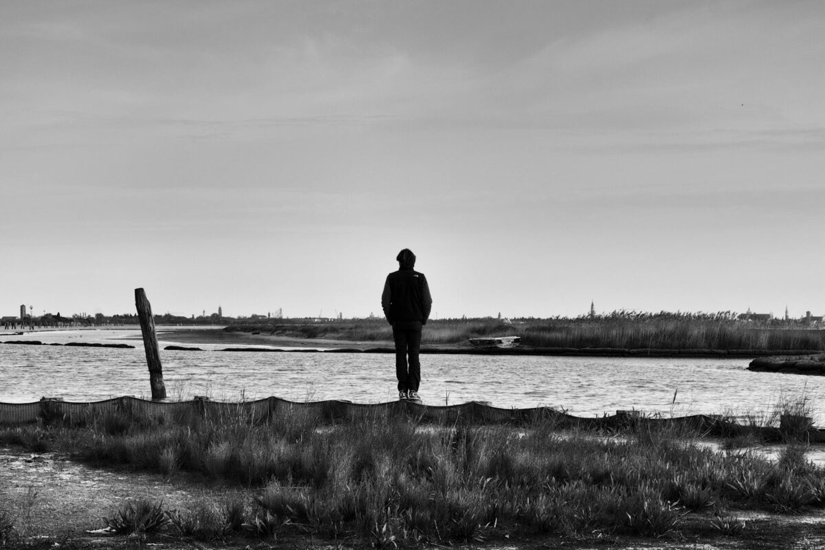 A person stands on a rural riverbank, embracing solitude and nature's serenity in black and white.