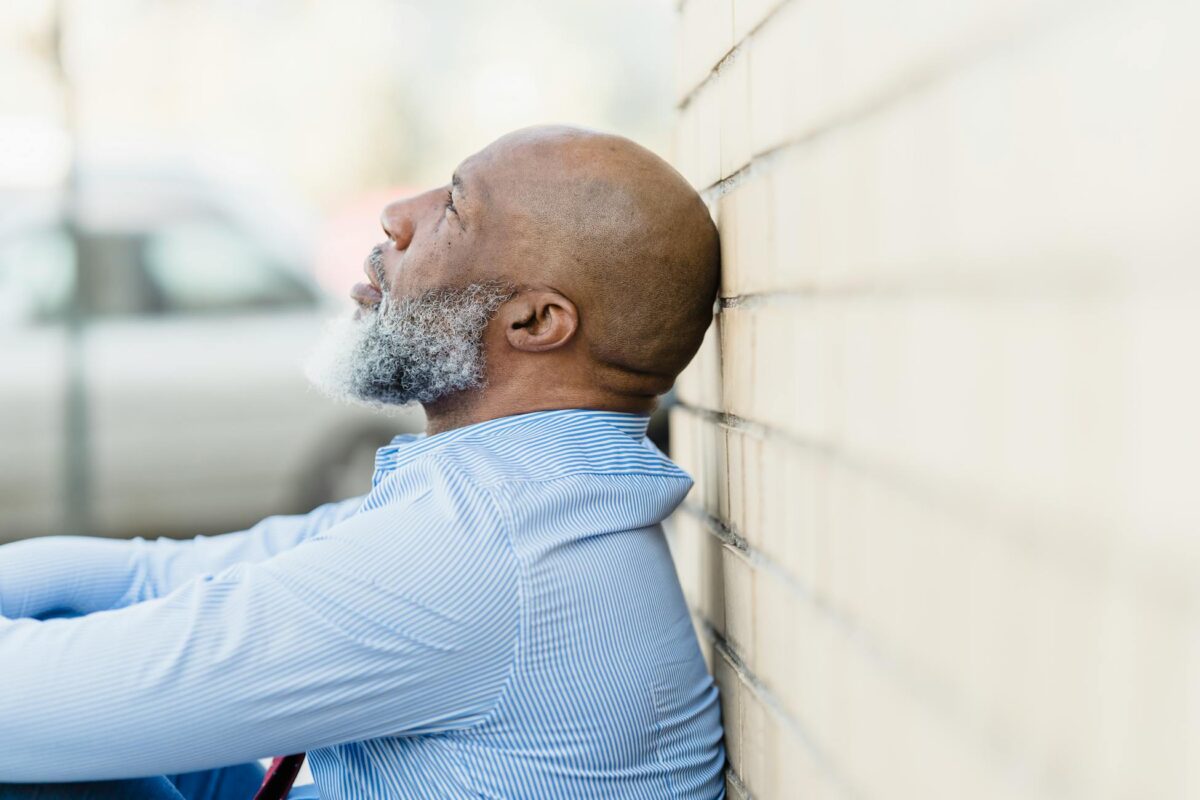 Side view of a thoughtful man with a beard leaning against an outdoor brick wall.