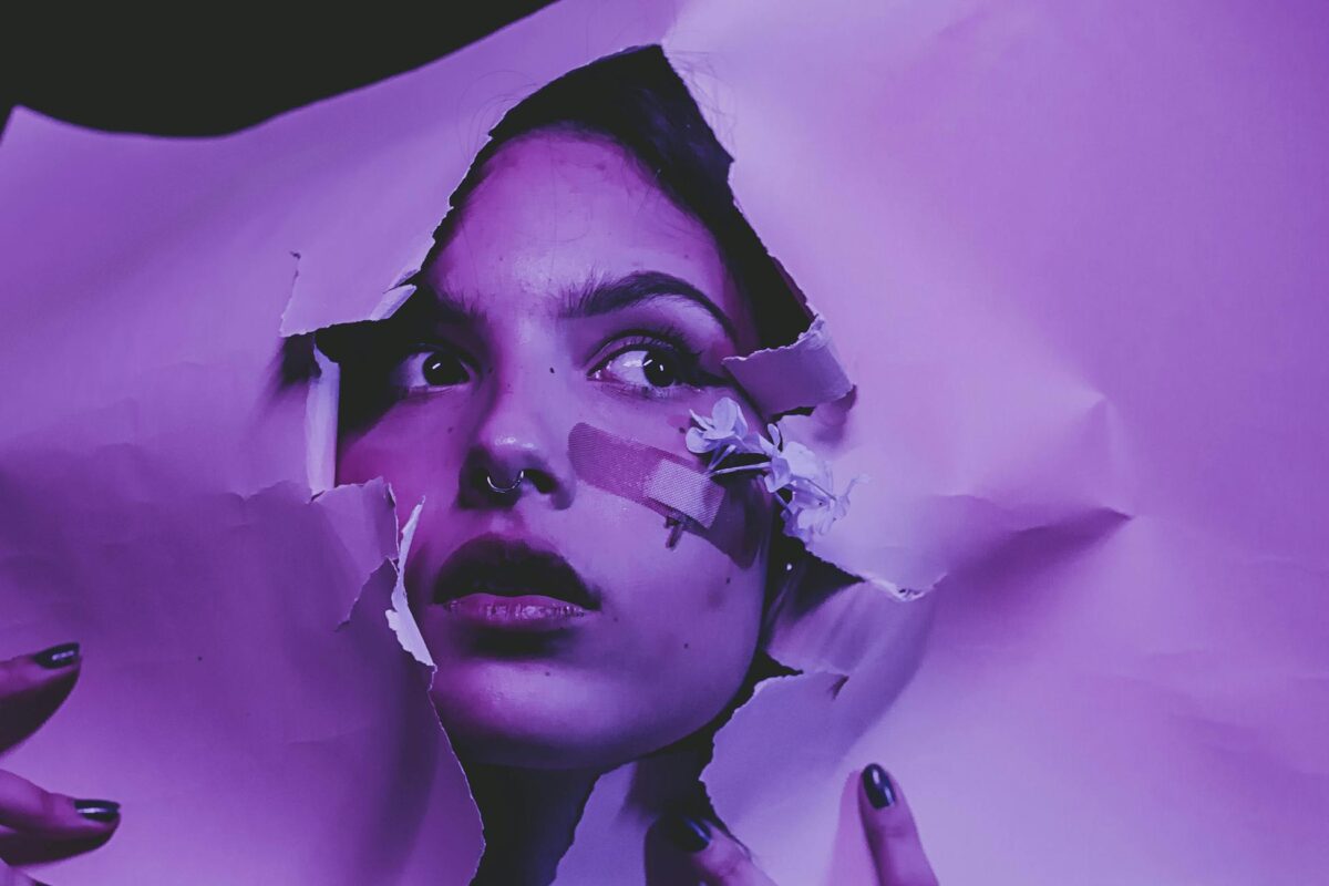 Artistic portrait of a woman looking sideways through torn purple paper, adorned with a band aid and flowers.