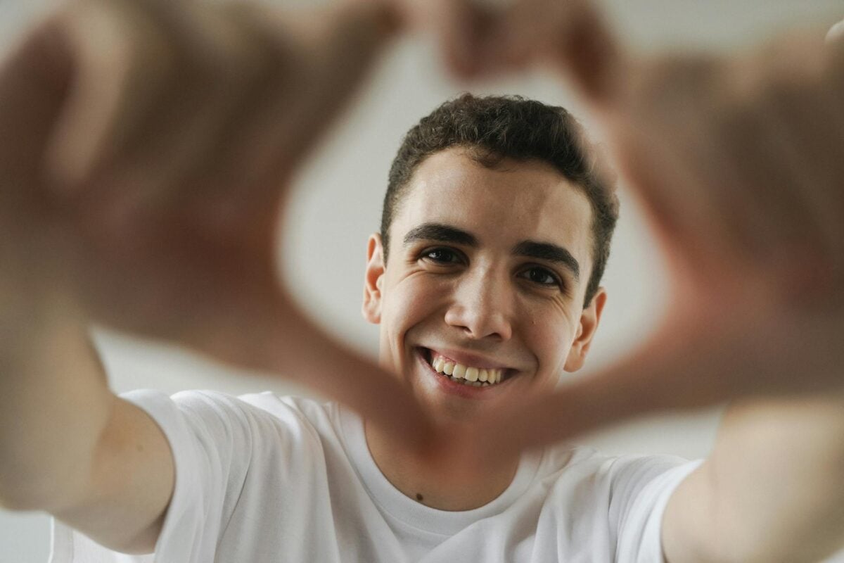 Close-up of a smiling young adult forming a heart shape with hands indoors.