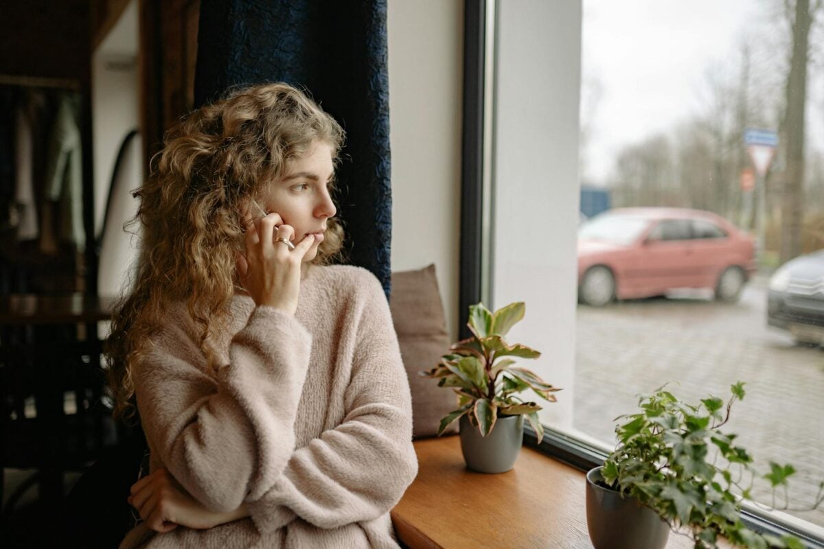 Woman in cozy sweater talking on a phone by the window, surrounded by houseplants, observing outdoors.