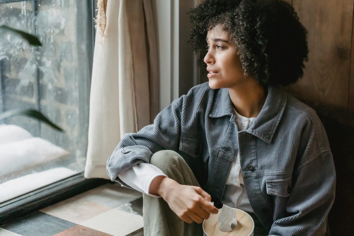 High angle of upset African American female suffering from loss and misfortune sitting on windowsill and looking away