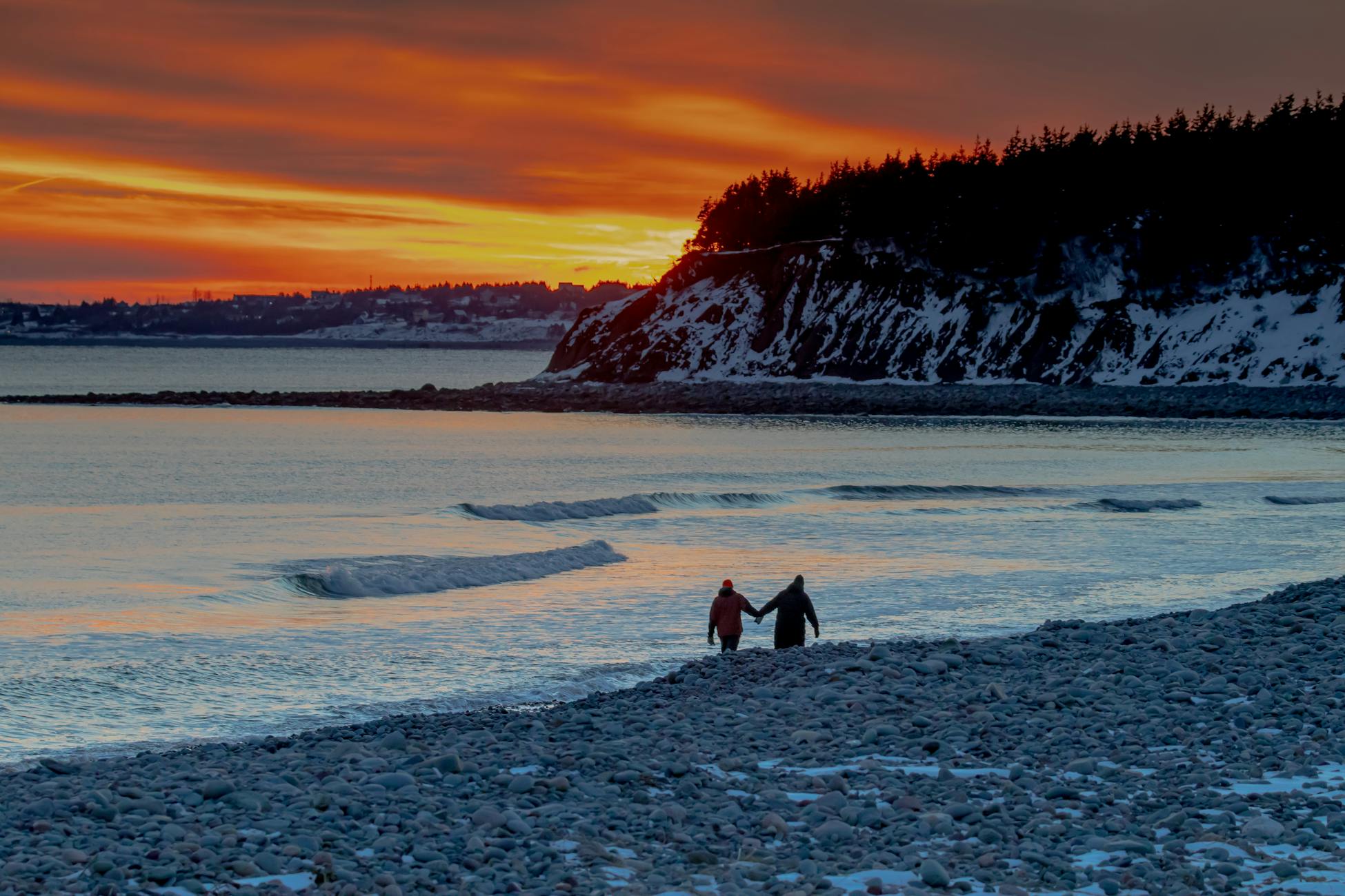 A couple holding hands on a rocky beach during a vibrant sunset by the ocean.