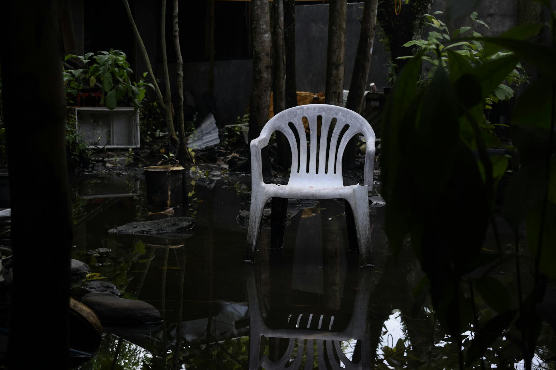 A white plastic chair reflecting in a waterlogged garden, surrounded by plants.
