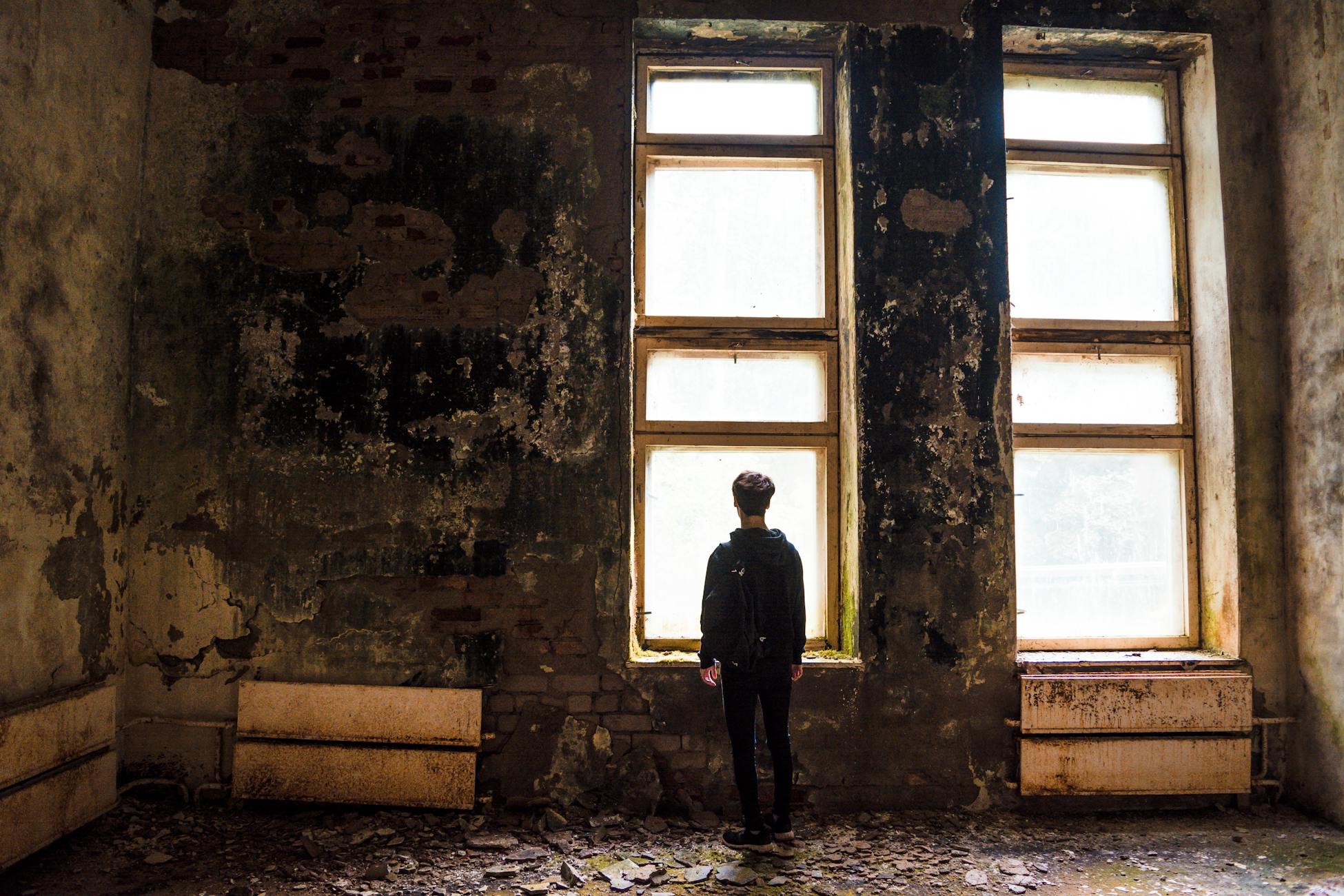 A solitary figure stands facing windows in a dilapidated urban setting, evoking themes of abandonment.