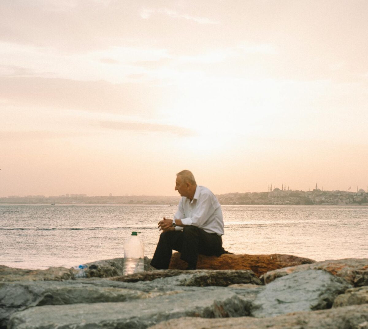 An elderly man sits alone on rocky shore, reflecting by the sea at sunset.