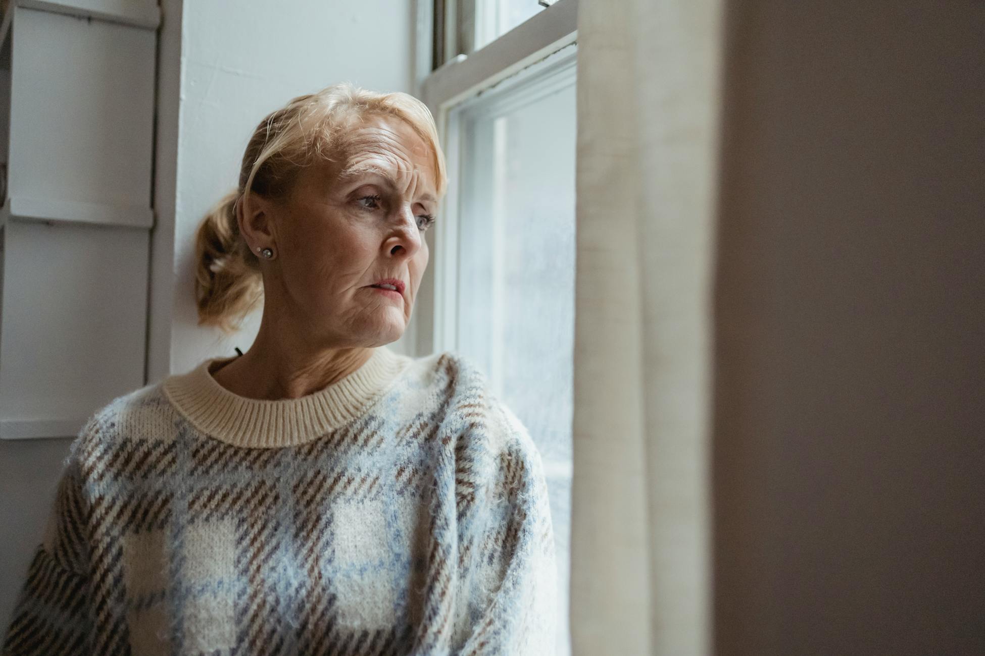 Contemplative senior female wearing warm sweater sitting on windowsill and looking away in sad thoughts