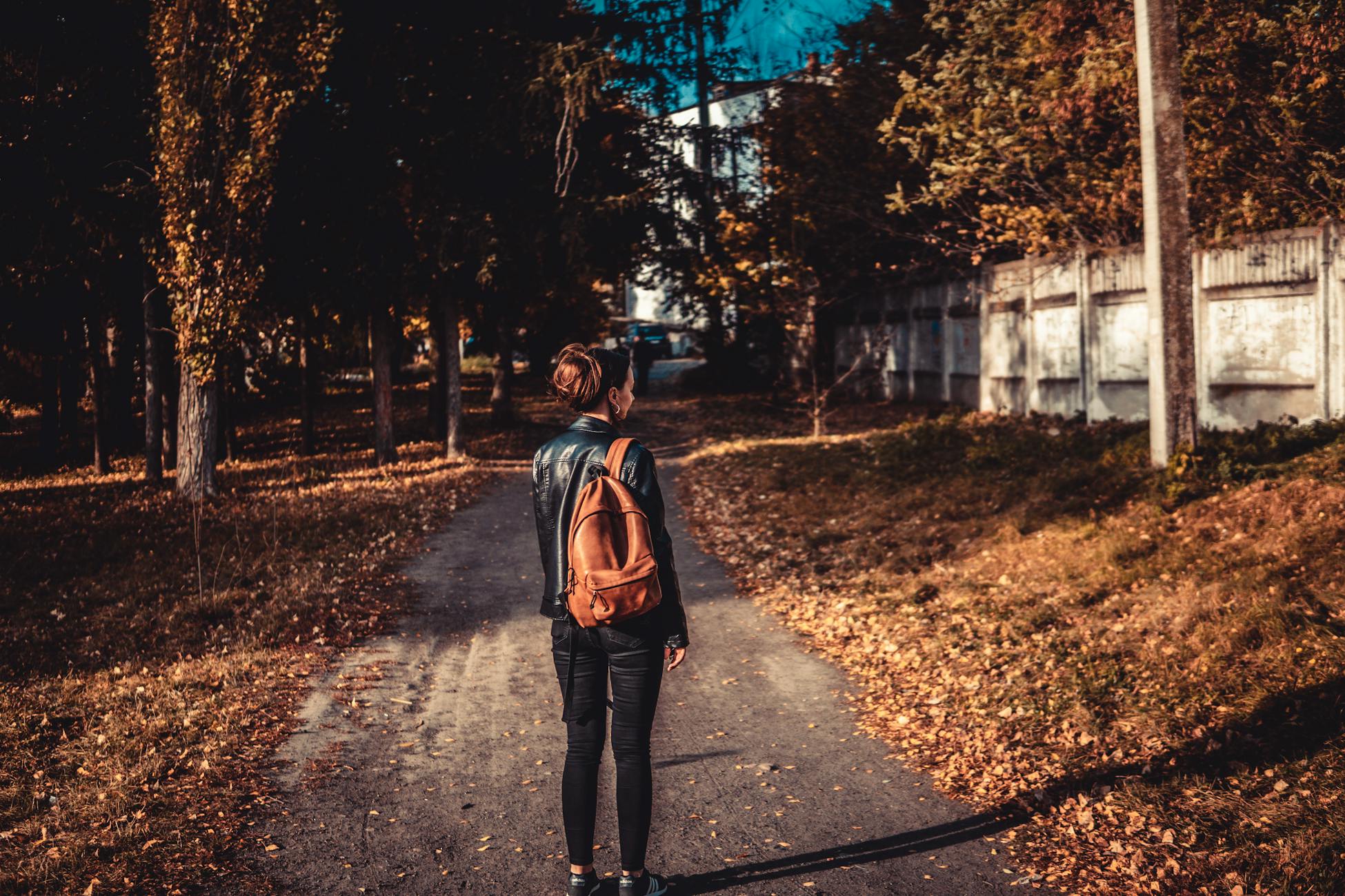 A woman with a brown backpack walks down a leafy path in the fall.