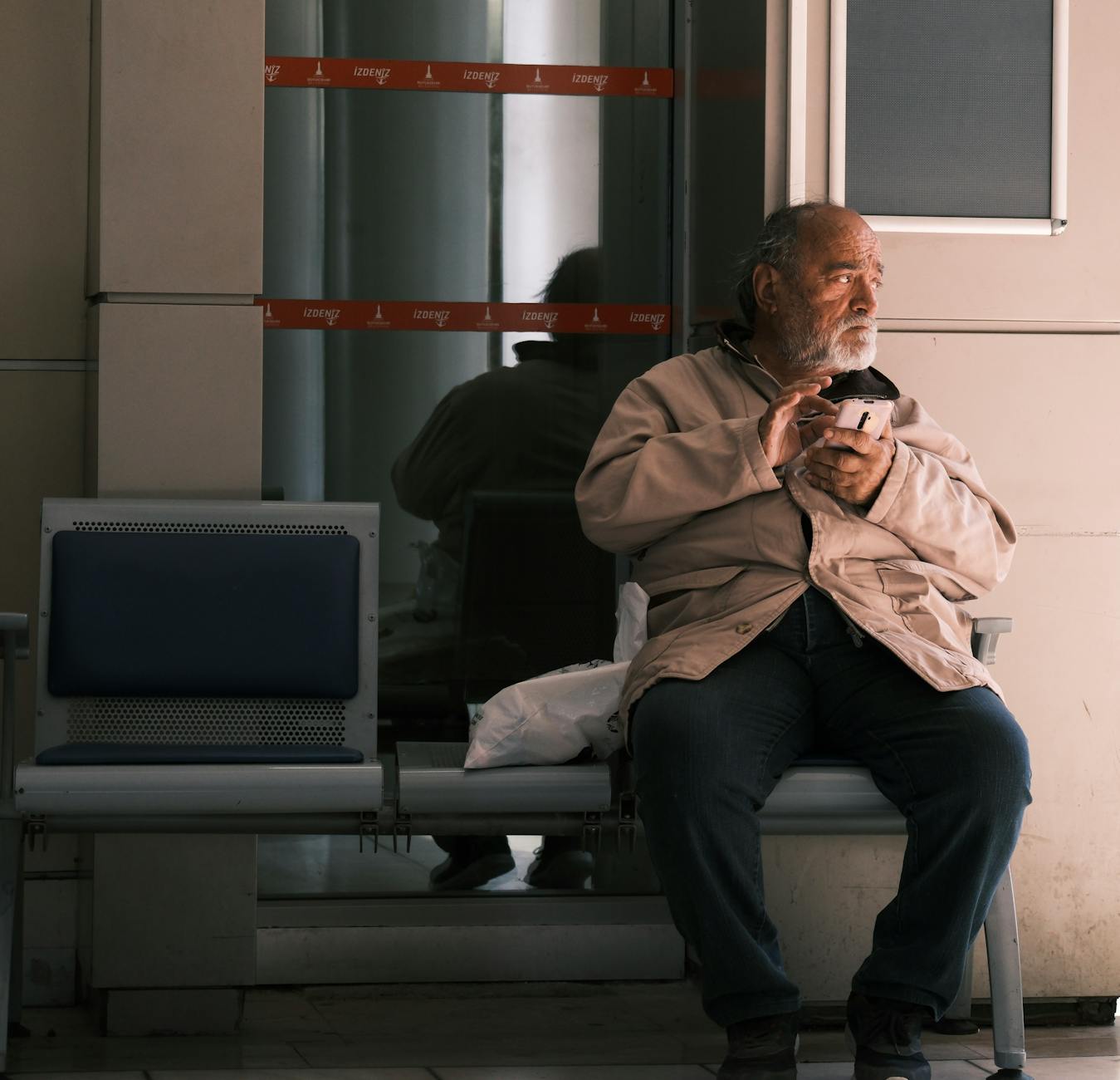 Elderly man holding a coffee cup, sitting alone on a bench indoors.