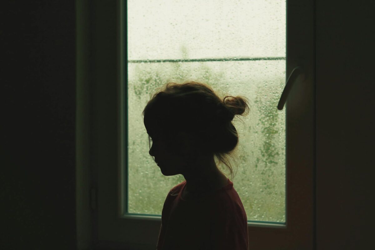 A child's silhouette stands by a rainy window, creating a nostalgic indoor scene.