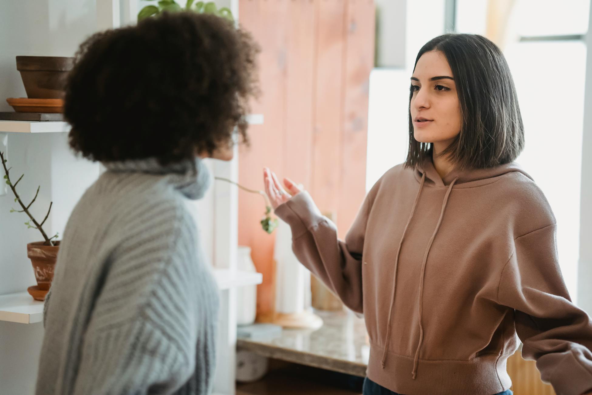 Serious woman in casual wear having argument with anonymous female with curly hair while standing in light room at home