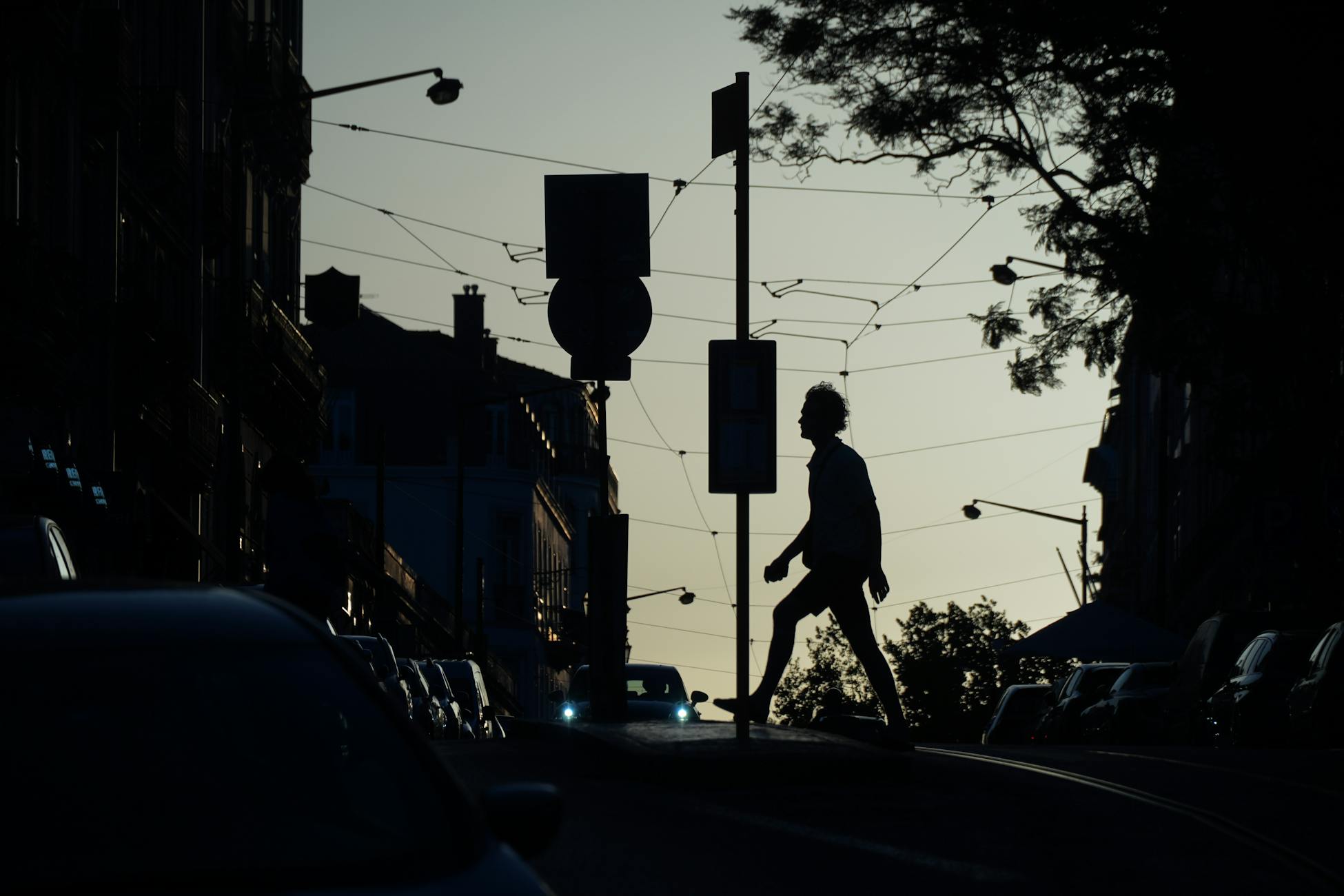 Silhouette of a person walking across a city street with cars and trees during sunset. Moody urban atmosphere.
