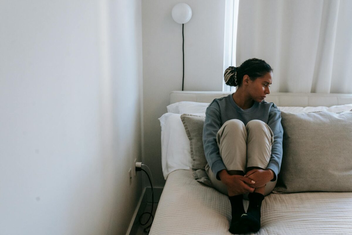 Young depressed ethnic female in casual wear looking away on soft bed in house