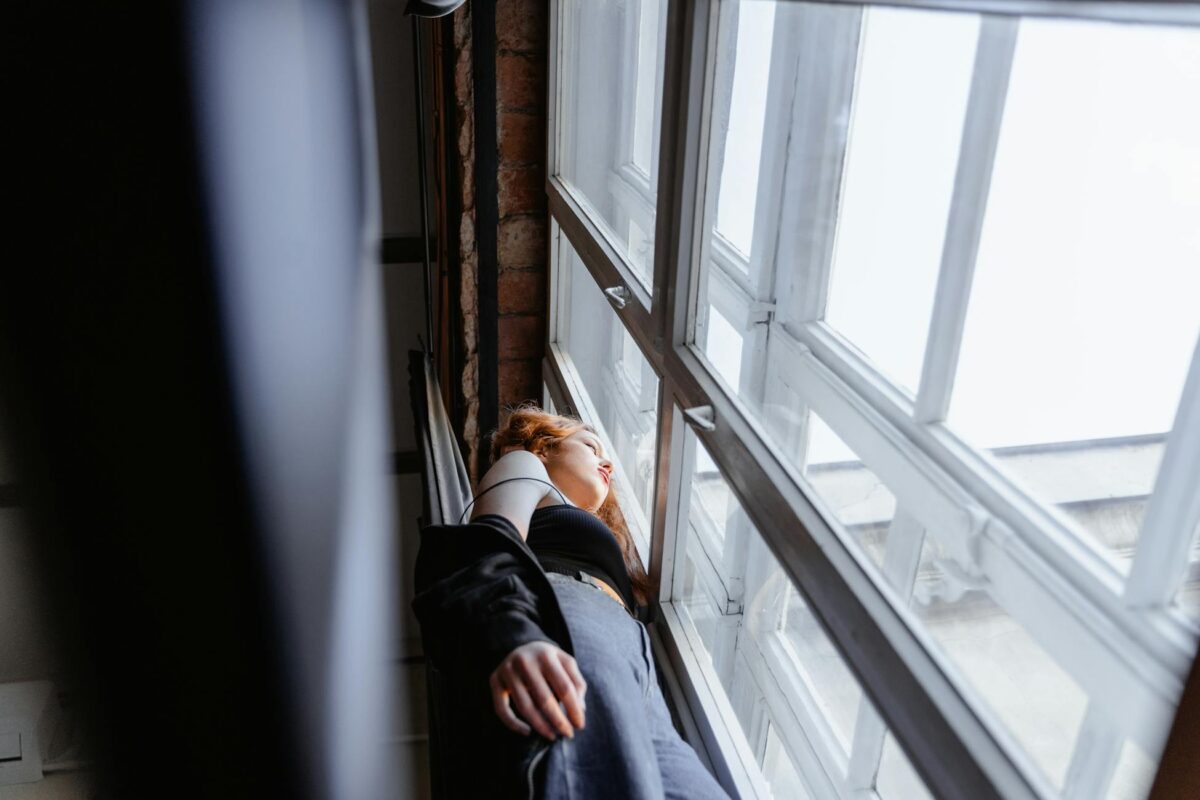 An adult woman leans against a window, lost in thought, indoors.